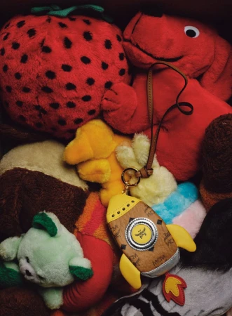 Close-up of stuffed toys including a red strawberry, dog, duck, bear and a rocket-shaped keychain from MCM in a pile.