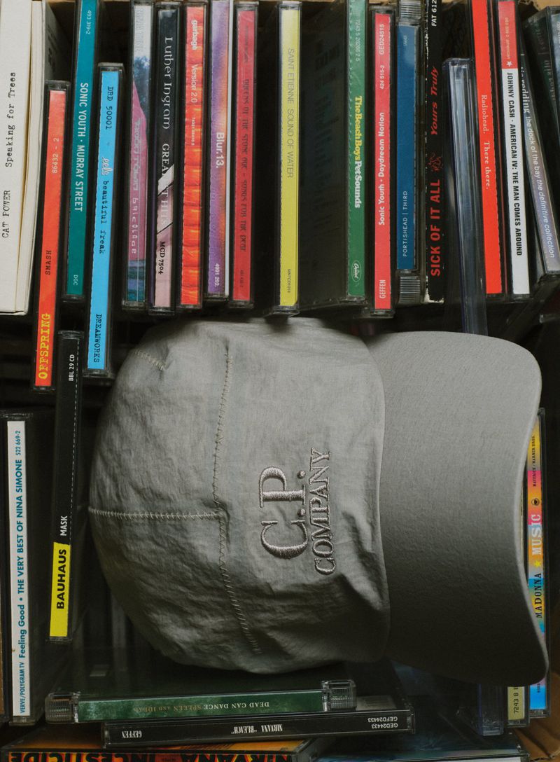 Grey C.P. Company cap displayed in front of a collection of colorful CD cases, creating a textured and musical backdrop.