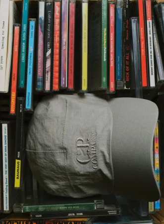 Grey C.P. Company cap displayed in front of a collection of colorful CD cases, creating a textured and musical backdrop.