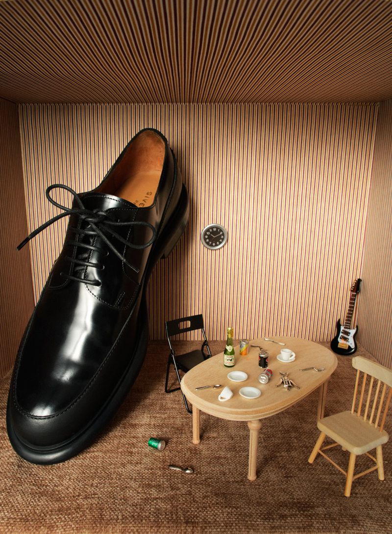 Artful still life featuring an oversized black dress shoe alongside miniature furniture, accessories, and decorations in a staged setting.