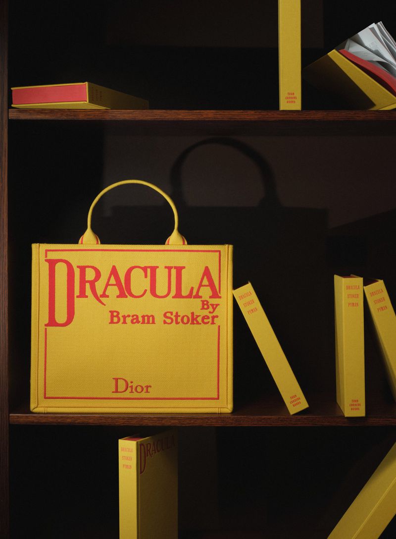 Yellow Dior book tote featuring Dracula by Bram Stoker on a dark wooden bookshelf with yellow books; a product shot emphasizing luxury fashion.