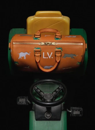 Fashion duffel bag with animal designs sits atop a green and yellow child's toy tractor in a studio setting against a black background.