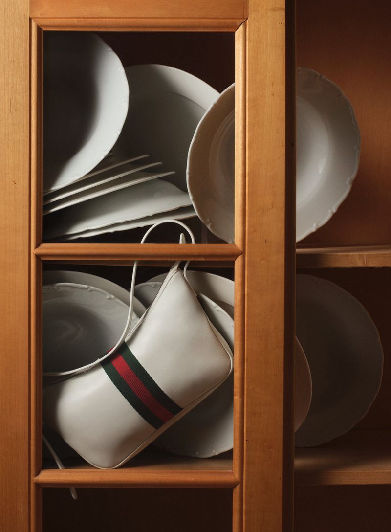 A white leather Gucci handbag with green and red stripe detail sits among stacked white plates inside of a wood cupboard with shelves.