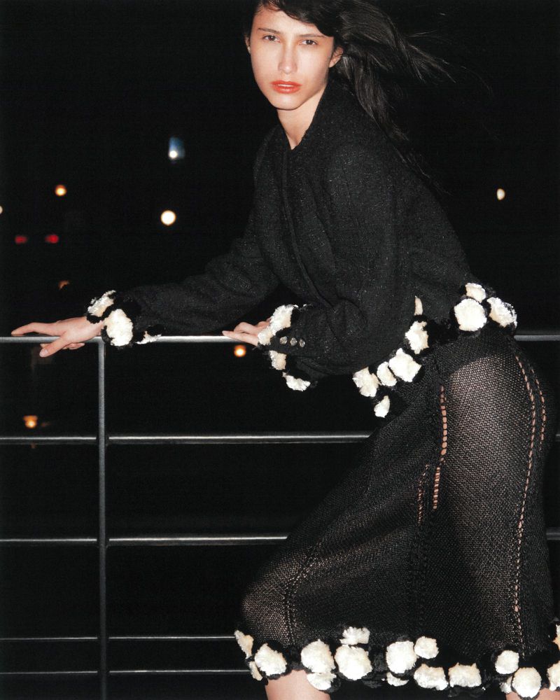 Elegant woman in dark jacket and sheer skirt with white floral accents poses at night against city lights for fashion editorial style shot.
