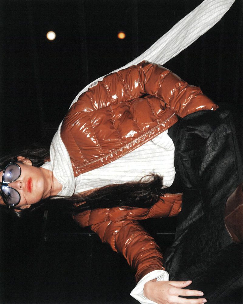 Fashionable woman in sunglasses and a brown puffer jacket poses indoors with dark background and soft, diffused lighting.