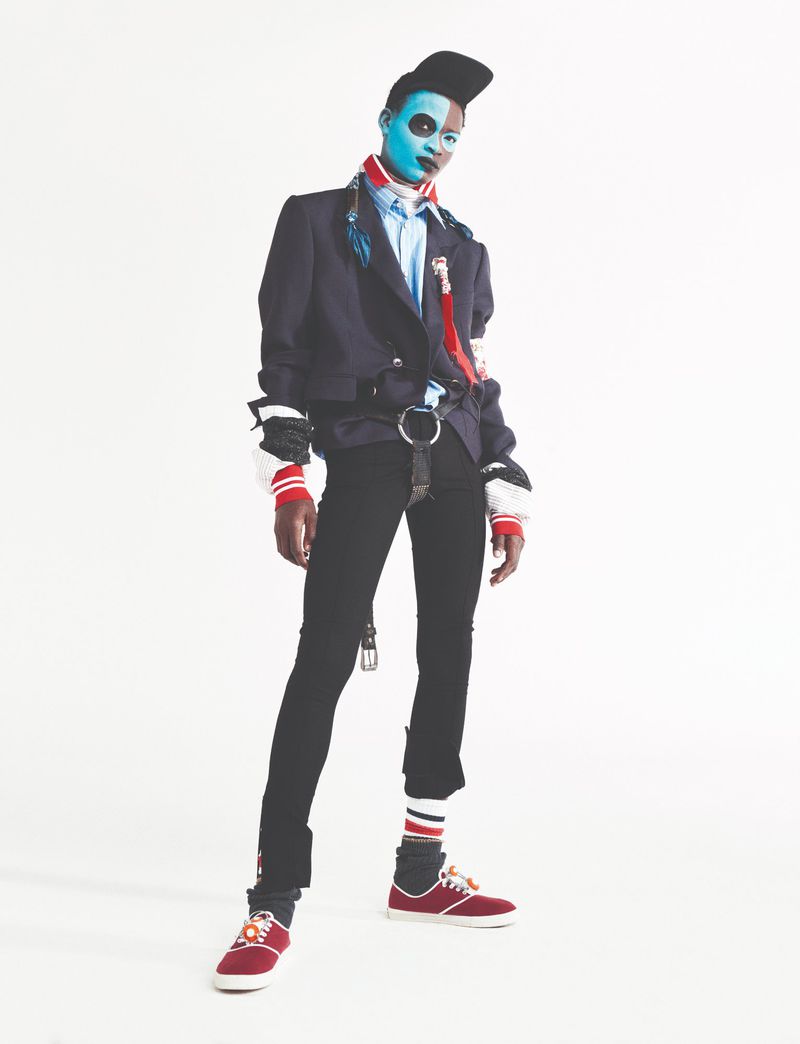 Full-length portrait of person with blue face paint, wearing a black jacket, black pants, and red sneakers against a white background.