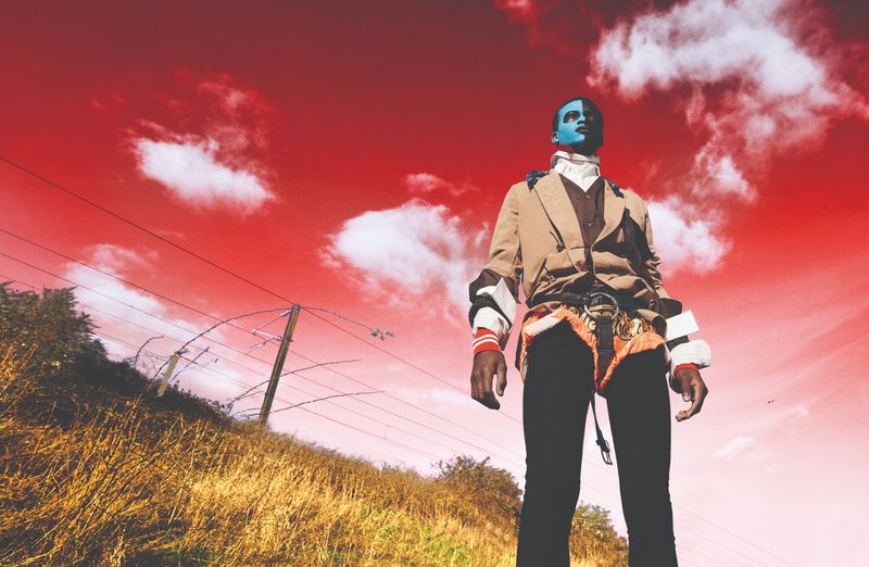 Adult with dramatic blue and white face paint stands in a beige jacket, black pants, and unique harness against a bright red sky.