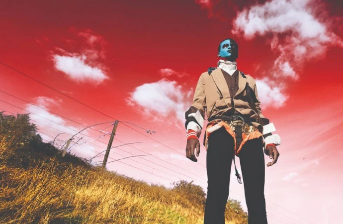 Adult with dramatic blue and white face paint stands in a beige jacket, black pants, and unique harness against a bright red sky.