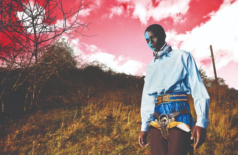 Portrait of an African-American adult male with blue face paint, wearing a blue shirt and decorative belt, outdoors in a field.