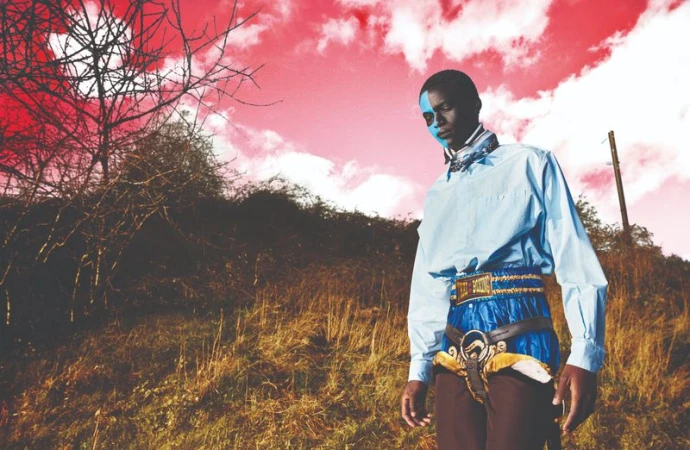 Portrait of an African-American adult male with blue face paint, wearing a blue shirt and decorative belt, outdoors in a field.