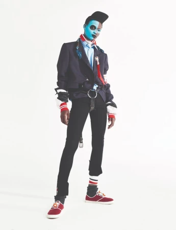 Model Seyi Boluwatife in dark blazer, black trousers, and cap with painted blue face standing against white background.
