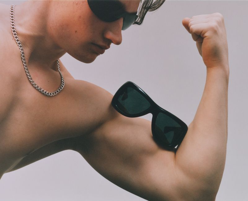 Close-up of a shirtless man with sunglasses flexing his bicep, which is being used as a stand for another pair of sunglasses.