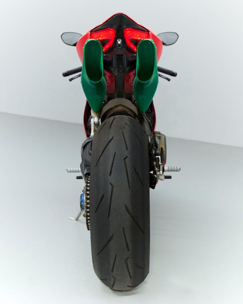 Back view of a red motorcycle with oversized green exhaust pipes against a white studio backdrop, highlighting the rear tire.