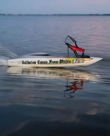 Powerboat races on the water with a woman's high-heel shoe mounted on the stern, reflecting in the water at sunset or sunrise.