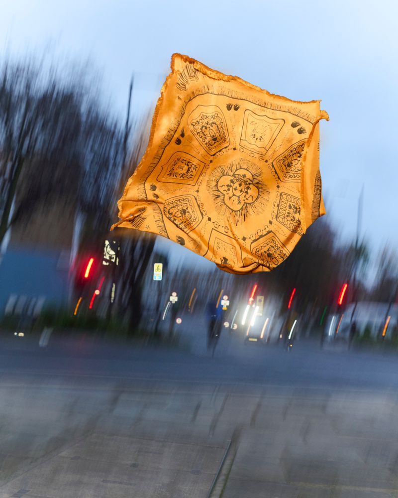 Golden patterned scarf levitates above an empty street with blurred motion and urban background, creating a surreal and artistic composition.