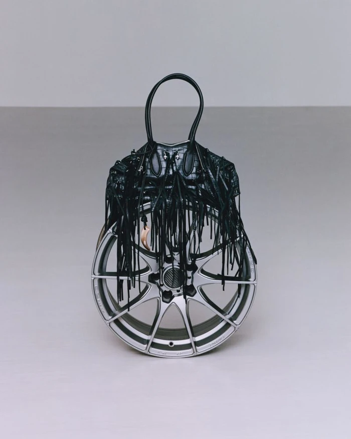 A black fringed handbag placed over a silver car wheel against a neutral grey studio background.