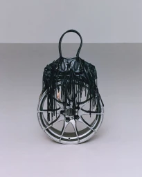 A black fringed handbag placed over a silver car wheel against a neutral grey studio background.