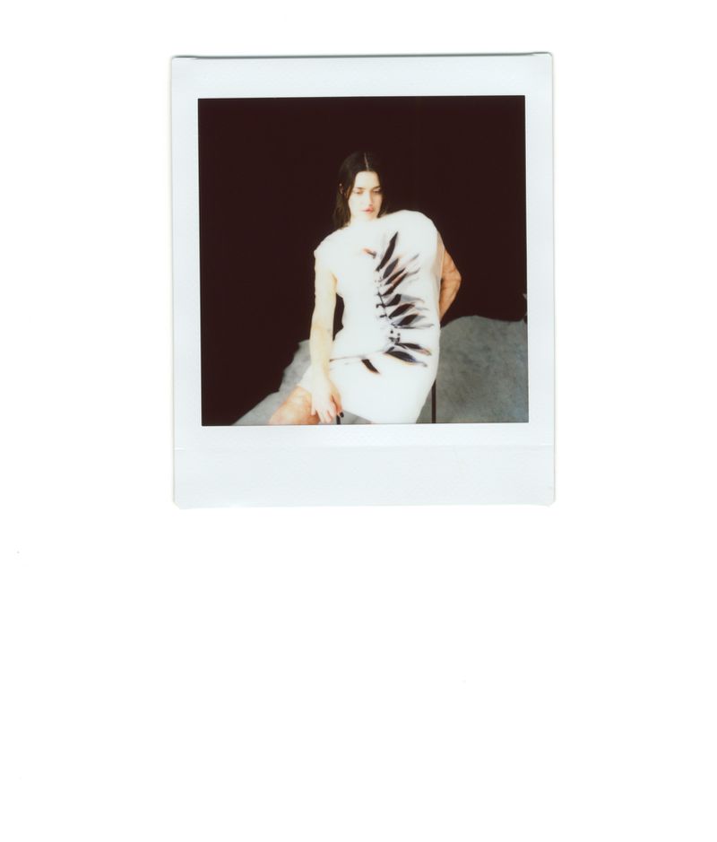 A woman sits with arms resting on a gray surface, dressed in a white dress with a black design, framed by a white border against a dark background.