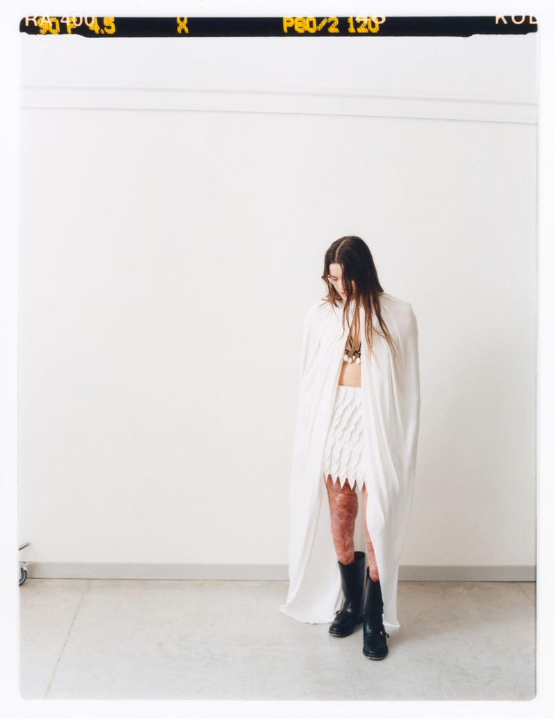 A person wearing white skirt and top with cape poses in front of a white wall with a film strip visible at the top.