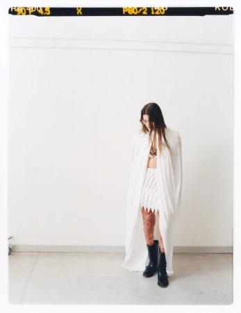 A person wearing white skirt and top with cape poses in front of a white wall with a film strip visible at the top.