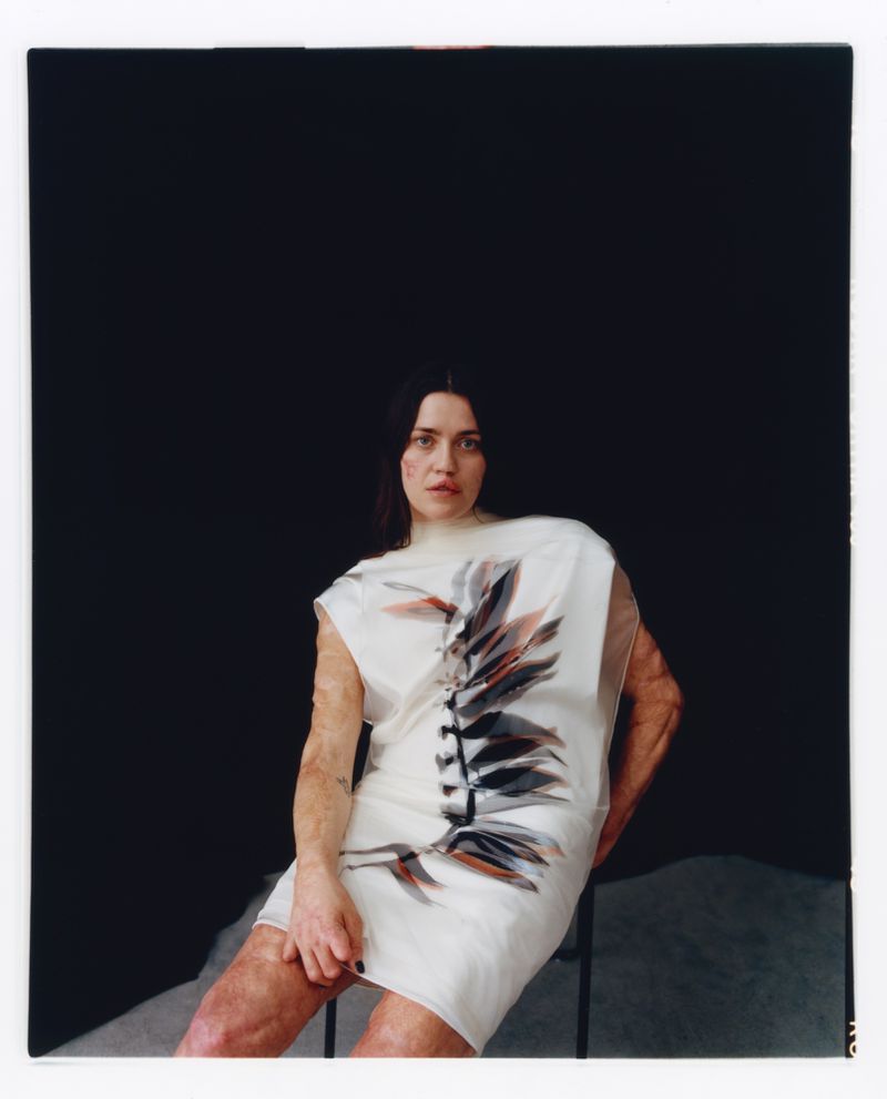 A woman with dark hair and a neutral expression, seated in a white dress with a plant print, poses against a dark gray backdrop.