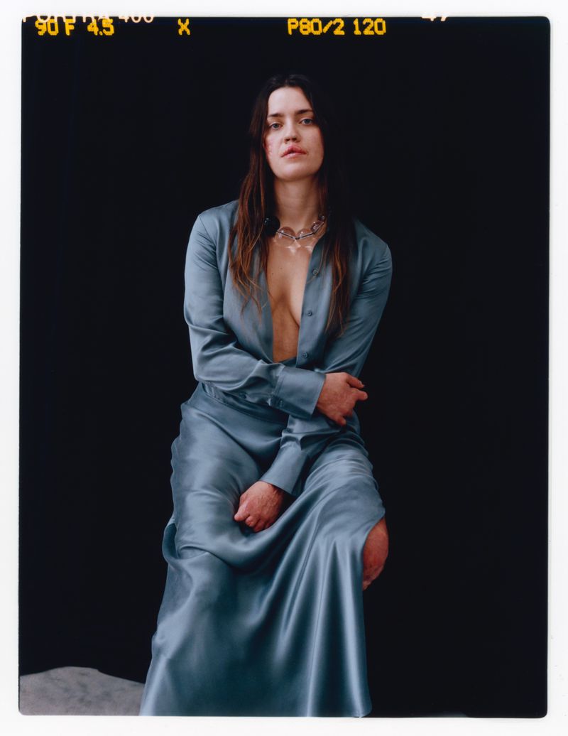 Portrait of a woman with long brown hair wearing a long-sleeved blue dress and silver necklace, sitting against a dark background.