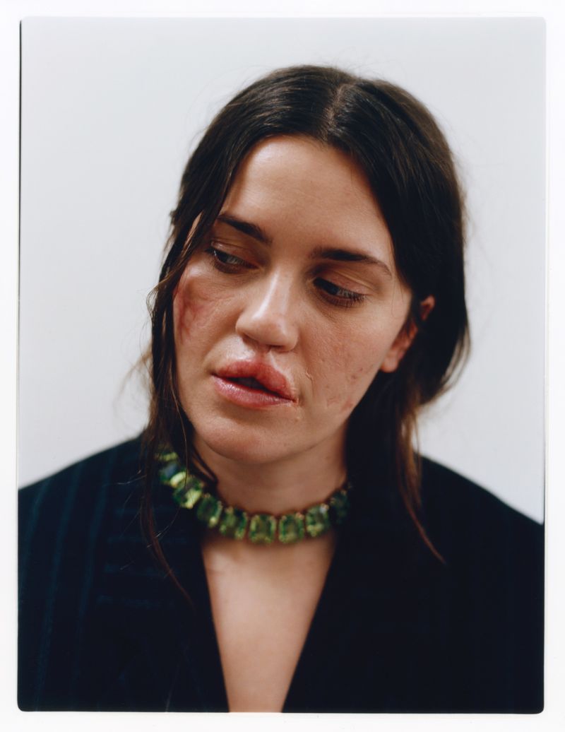 Close-up portrait of a woman with dark hair and a striking green gemstone necklace against a clean white background in a studio setting.