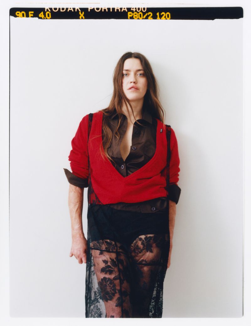 A woman with long brown hair poses indoors wearing a red sweater, brown blouse, and a black lace skirt with a white wall backdrop.