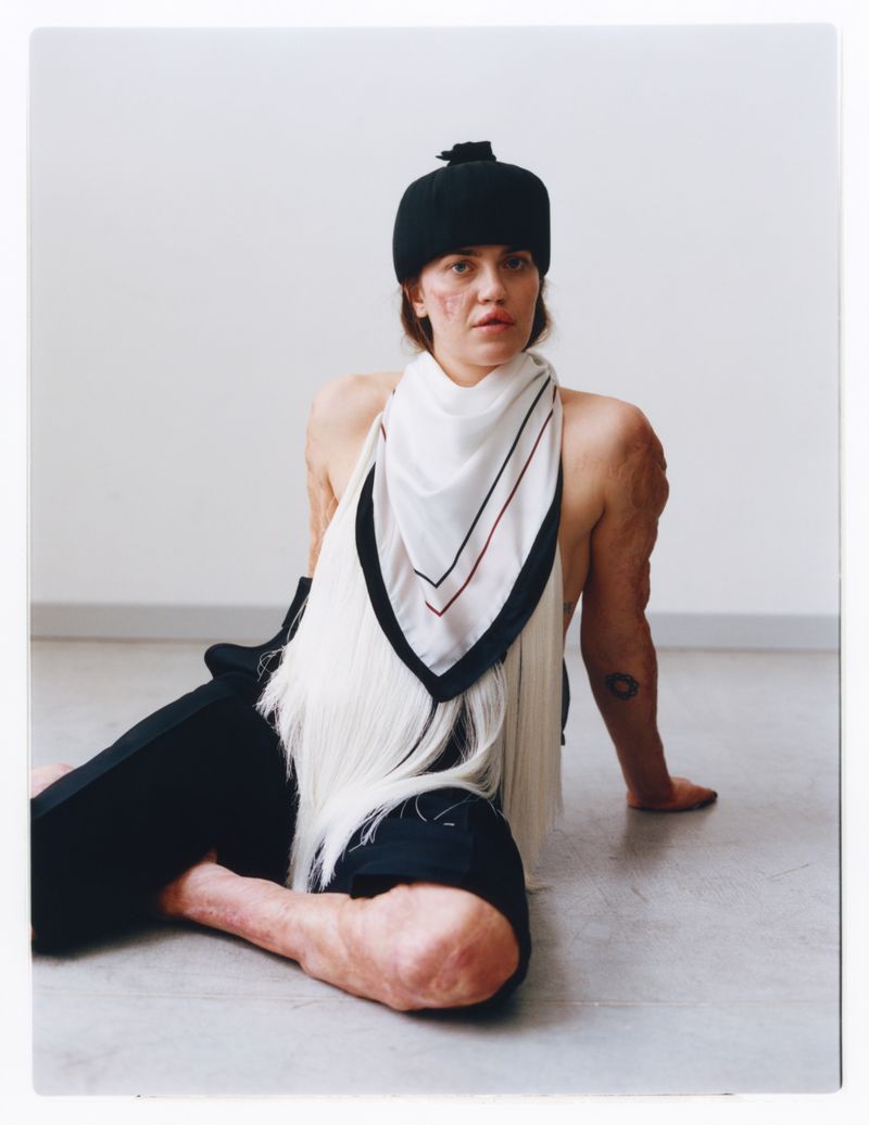 A woman in a black cap and scarf poses indoors. The image is a waist-up portrait with a neutral background, emphasising fashion and beauty.