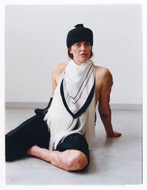 A woman in a black cap and scarf poses indoors. The image is a waist-up portrait with a neutral background, emphasising fashion and beauty.