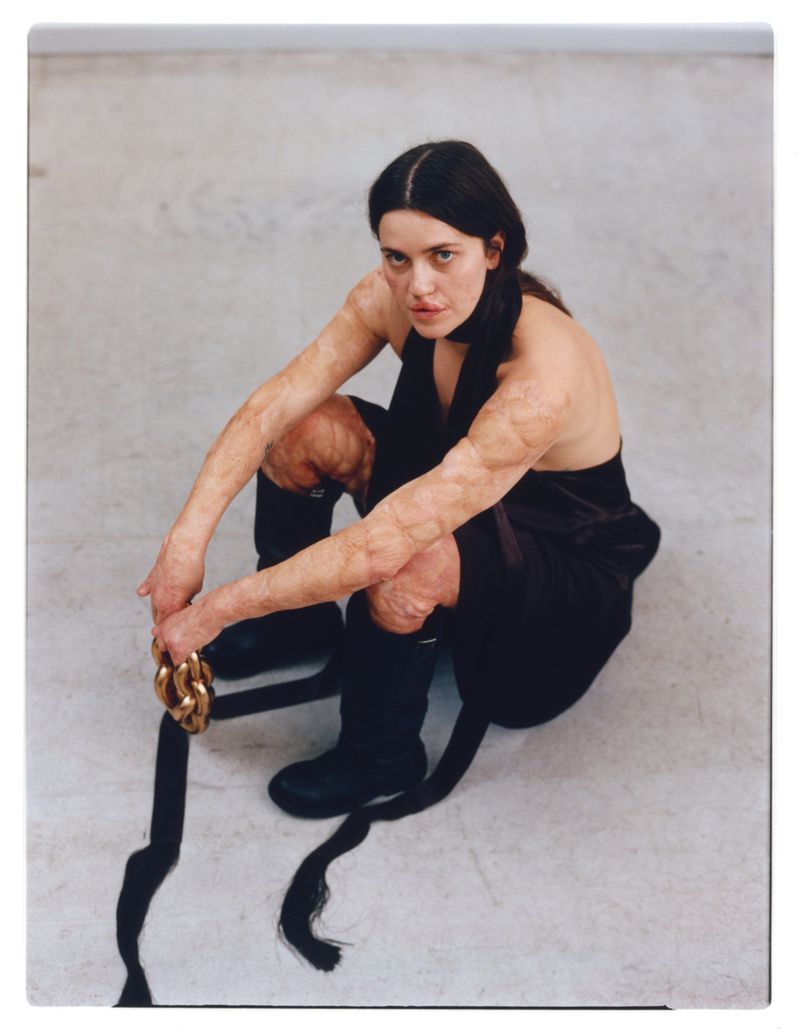 An adult woman in a dark dress poses indoors, kneeling on a concrete floor while holding a gold chain and wearing dark-colored boots.