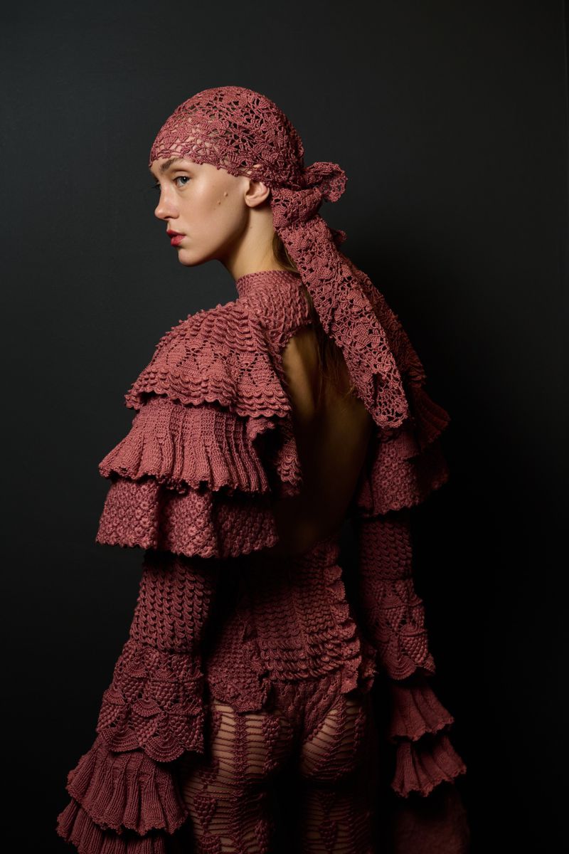 Model in mauve crocheted ruffled garment and headscarf posing against a dark background for J.Salinas AW26 runway editorial.