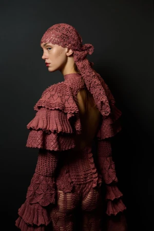 Model in mauve crocheted ruffled garment and headscarf posing against a dark background for J.Salinas AW26 runway editorial.