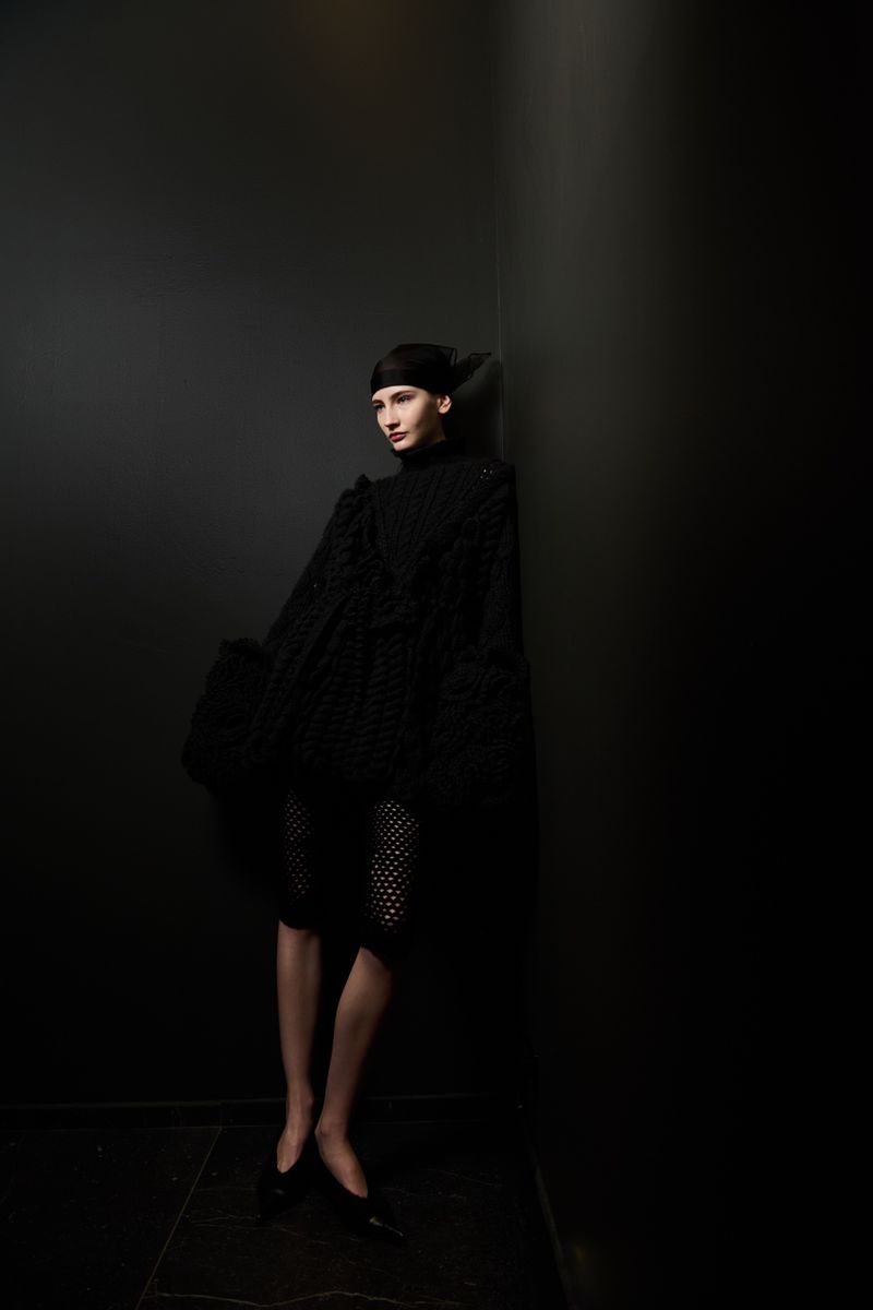 A solitary female model leans against a dark gray wall in a textured black dress, complemented by matching flats and head covering.