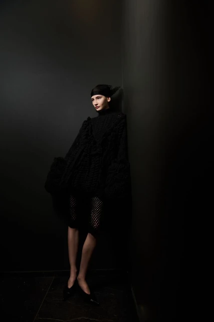 A solitary female model leans against a dark gray wall in a textured black dress, complemented by matching flats and head covering.