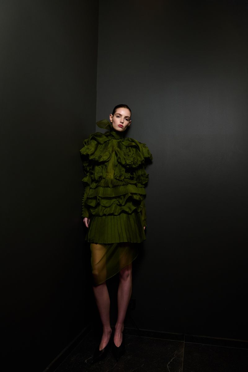 A woman posing in an olive green ruffled dress against a dark grey wall, creating a dramatic fashion portrait with high contrast.