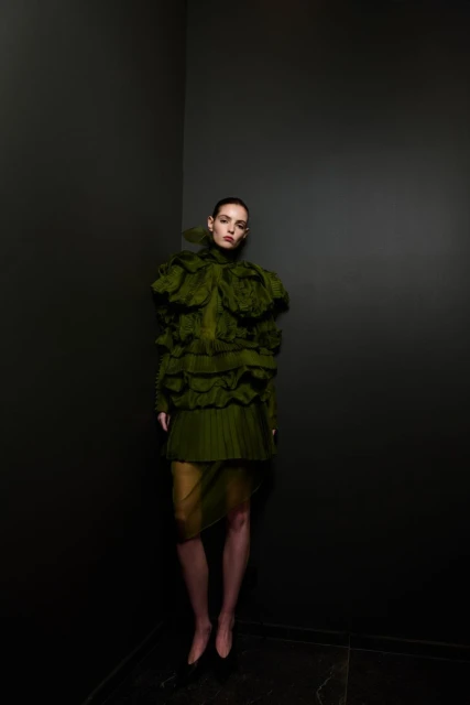 A woman posing in an olive green ruffled dress against a dark grey wall, creating a dramatic fashion portrait with high contrast.