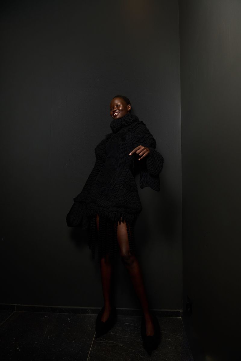 A Black female model in a black turtleneck dress poses in a corner with black walls, showing off an editorial fashion look and style.