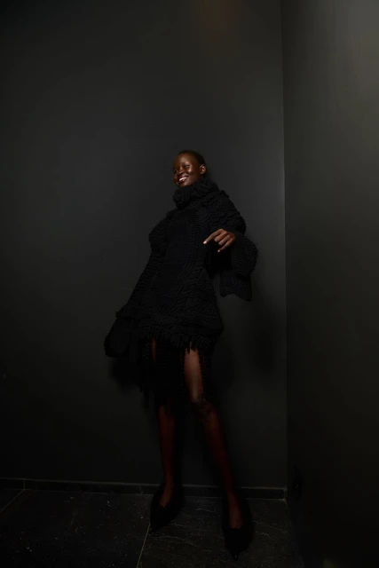 A Black female model in a black turtleneck dress poses in a corner with black walls, showing off an editorial fashion look and style.