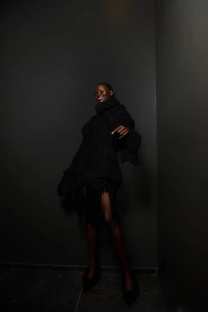 A Black female model in a black turtleneck dress poses in a corner with black walls, showing off an editorial fashion look and style.