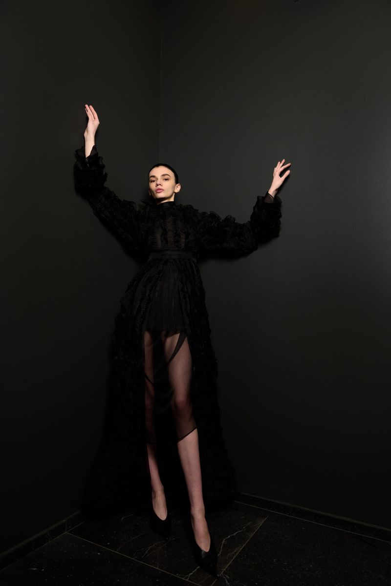 Fashion photo of a woman in a black textured dress with raised arms, posing against a dark gray wall in an indoor studio setting.