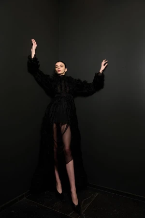 Fashion photo of a woman in a black textured dress with raised arms, posing against a dark gray wall in an indoor studio setting.