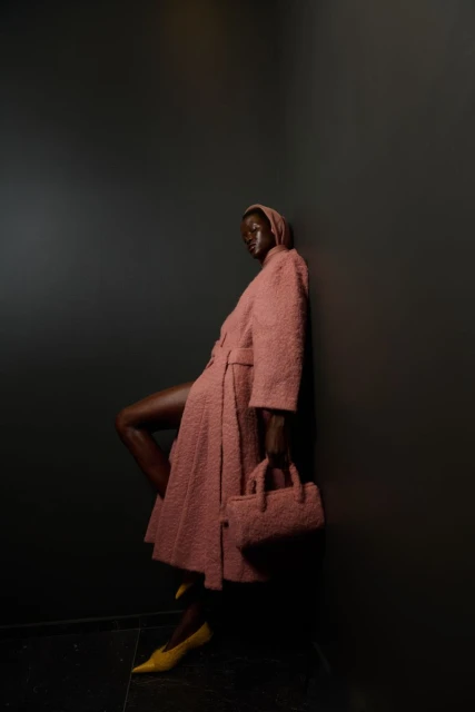 Dark-skinned woman in a hooded rose overcoat leans against a dark wall holding a matching purse, complemented by yellow heels.