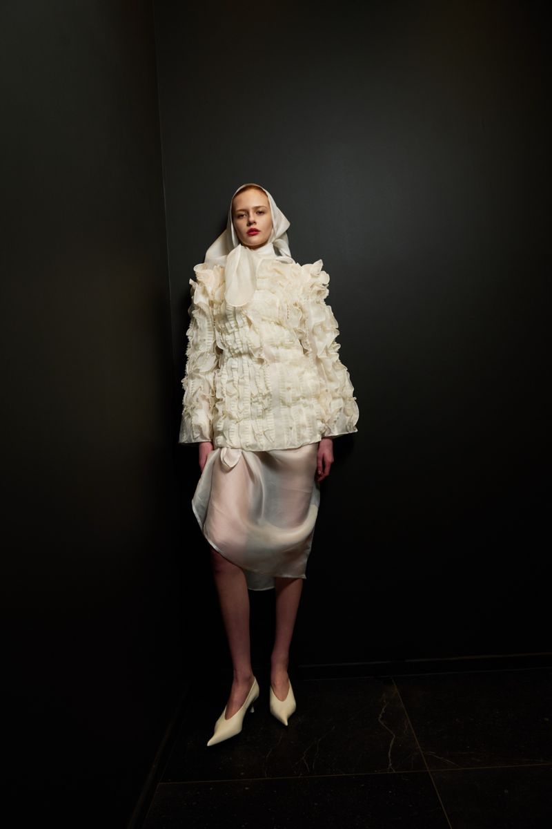 A woman models an avant-garde ruffled jacket and skirt with a headscarf against a dark grey wall and black tile floor indoors.
