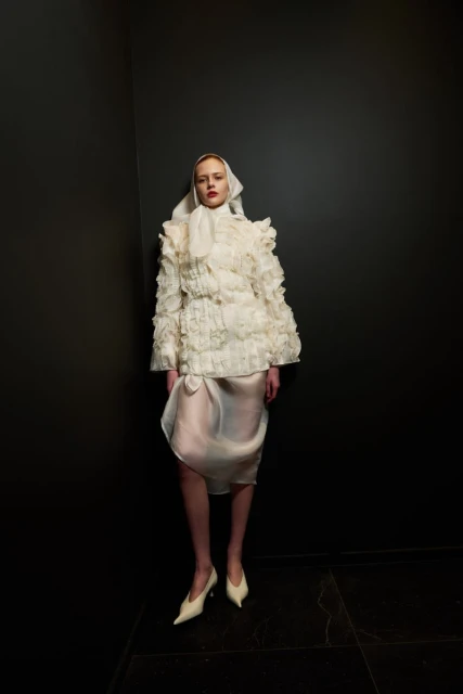 A woman models an avant-garde ruffled jacket and skirt with a headscarf against a dark grey wall and black tile floor indoors.