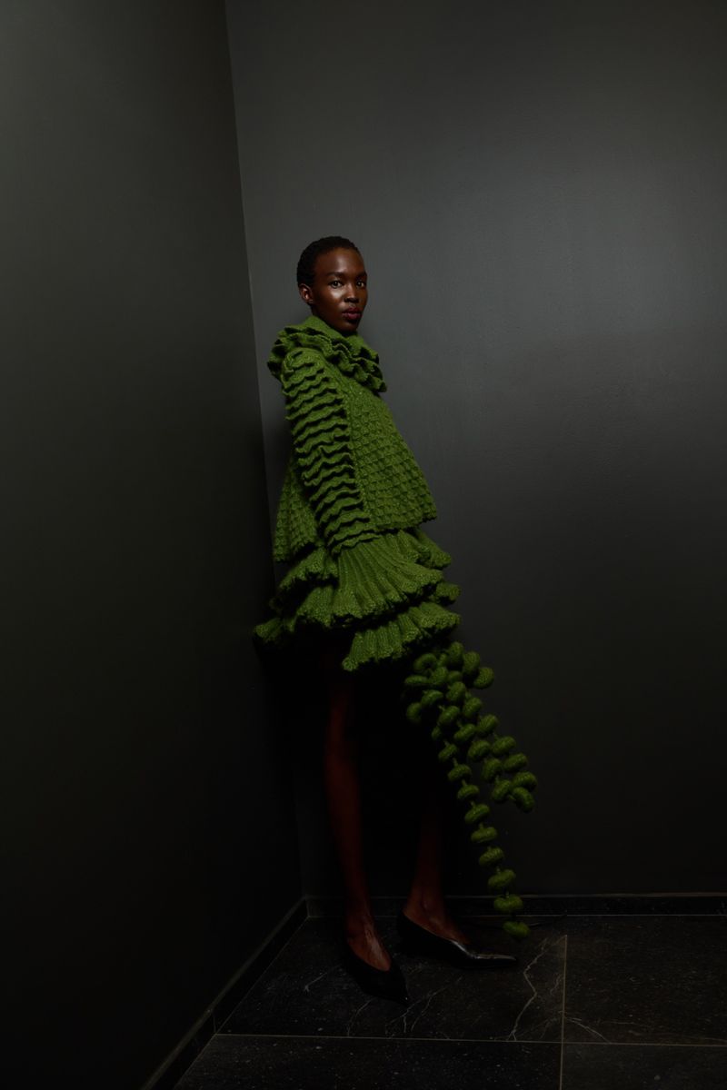 Black woman in a green, tiered, ruffled dress with black stripes poses in a grey corner with tiled floors, fashion photoshoot.