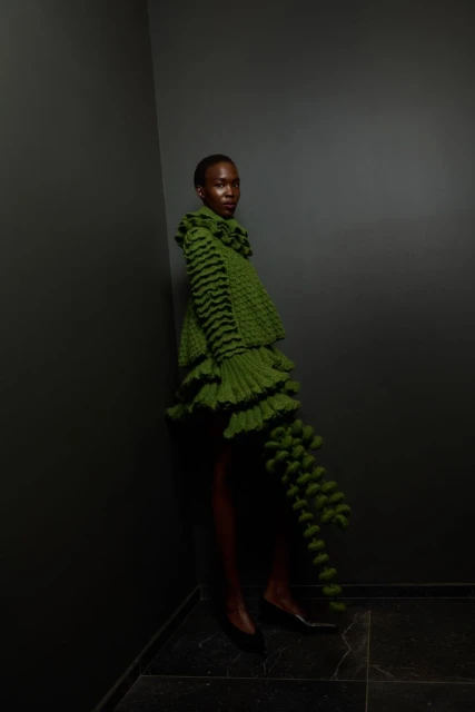 Black woman in a green, tiered, ruffled dress with black stripes poses in a grey corner with tiled floors, fashion photoshoot.