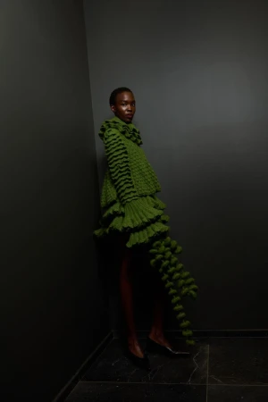 Black woman in a green, tiered, ruffled dress with black stripes poses in a grey corner with tiled floors, fashion photoshoot.