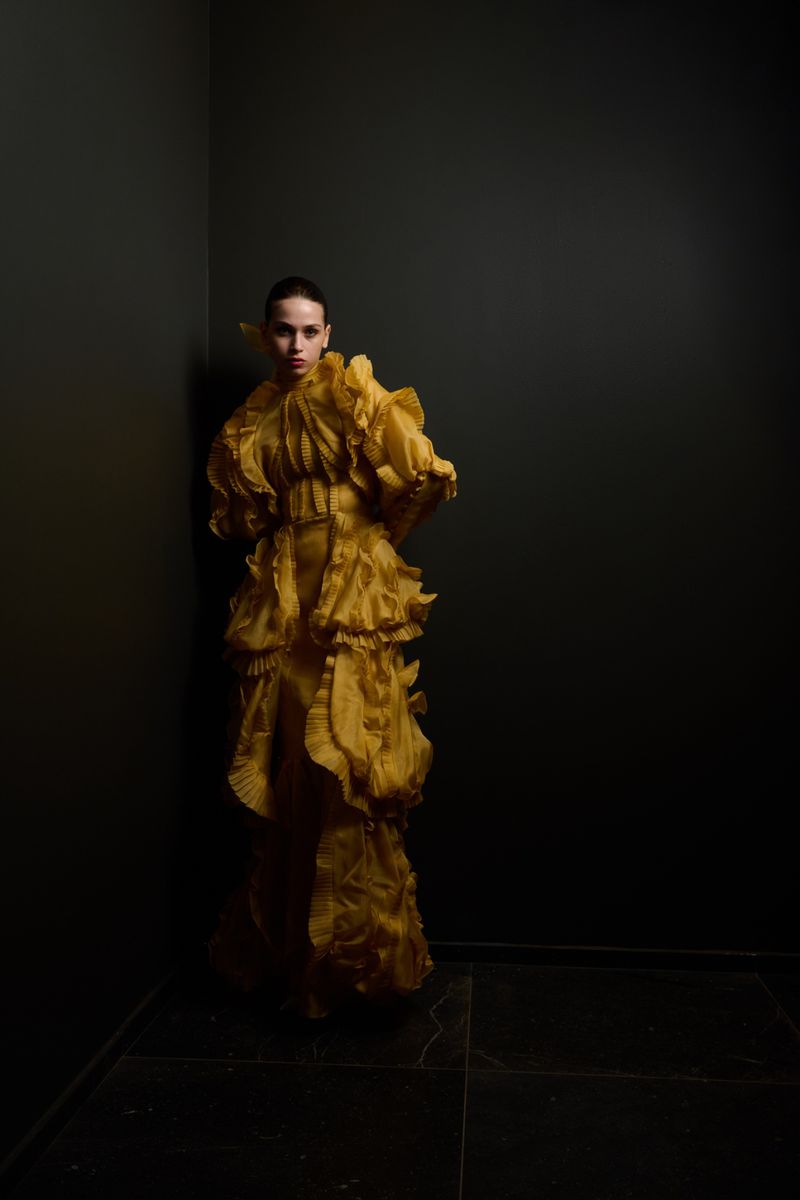 A stylish adult female in a yellow ruffled gown stands in a dark corner. Editorial fashion shot with a focus on color and texture.