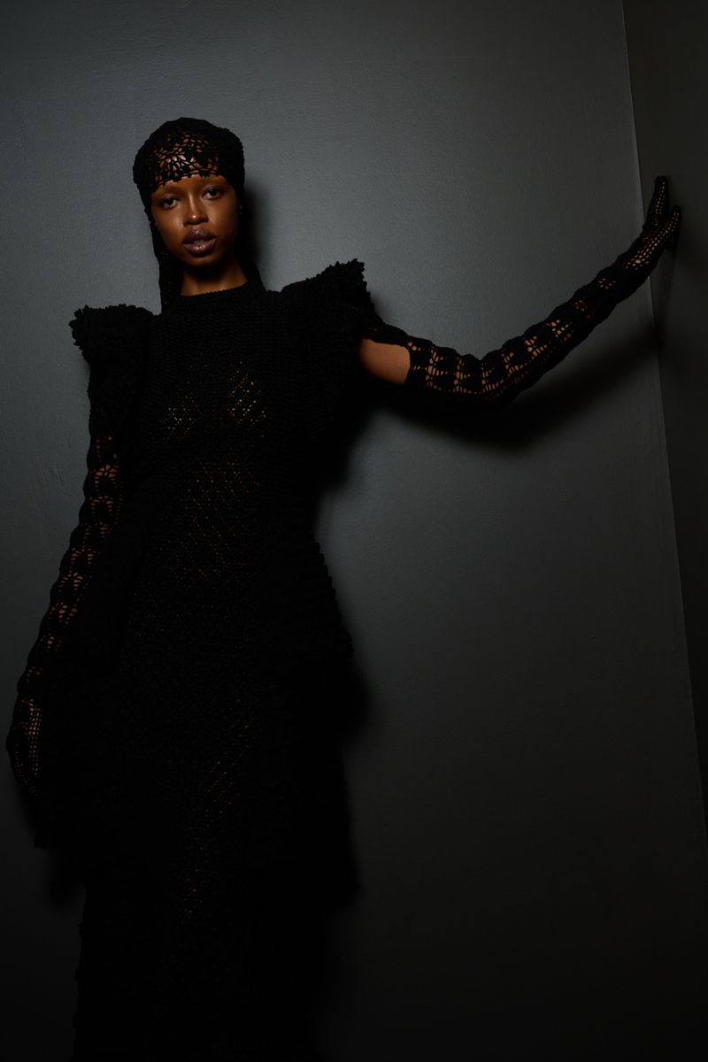 Portrait of a Black woman in a black lace dress and long gloves, posing against a dark gray wall in a fashion studio.